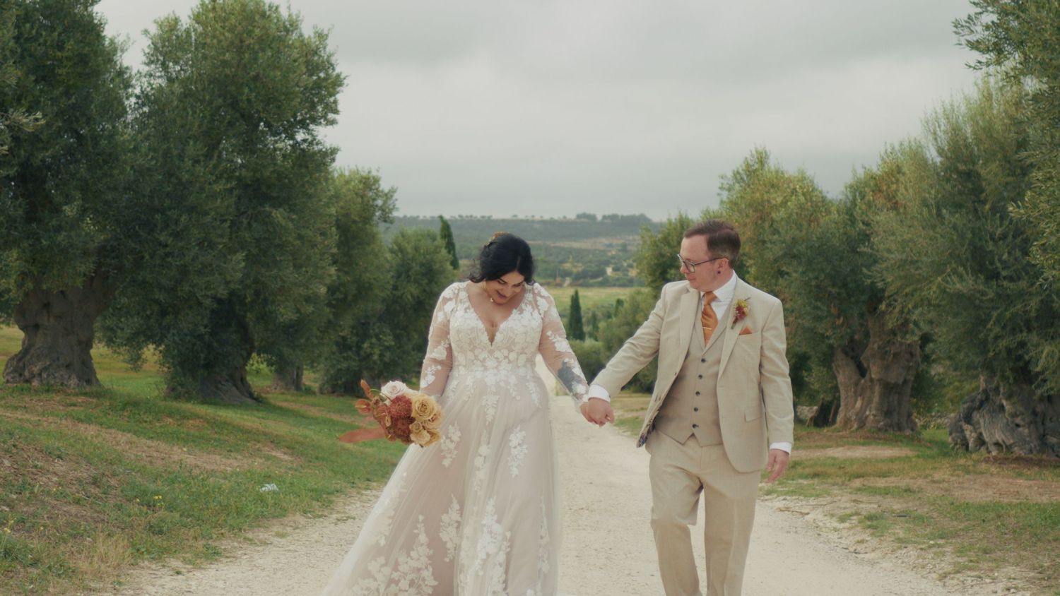 Bride and groom walking hand in hand along a scenic dirt path lined with olive trees on their wedding day.
