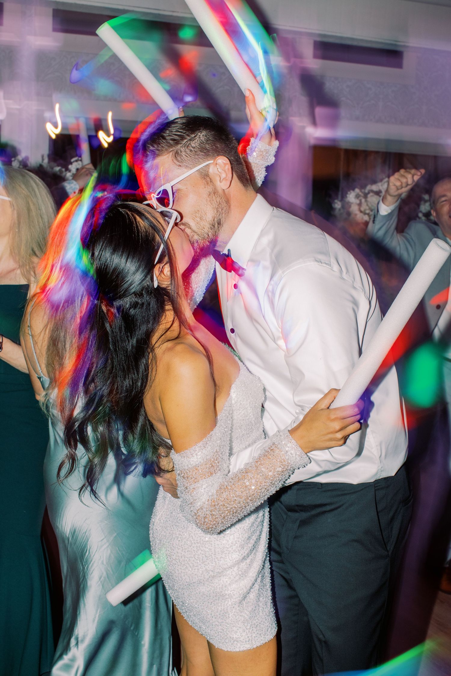 Couple kissing on a colorful dance floor with glowing foam sticks at a lively wedding reception party.