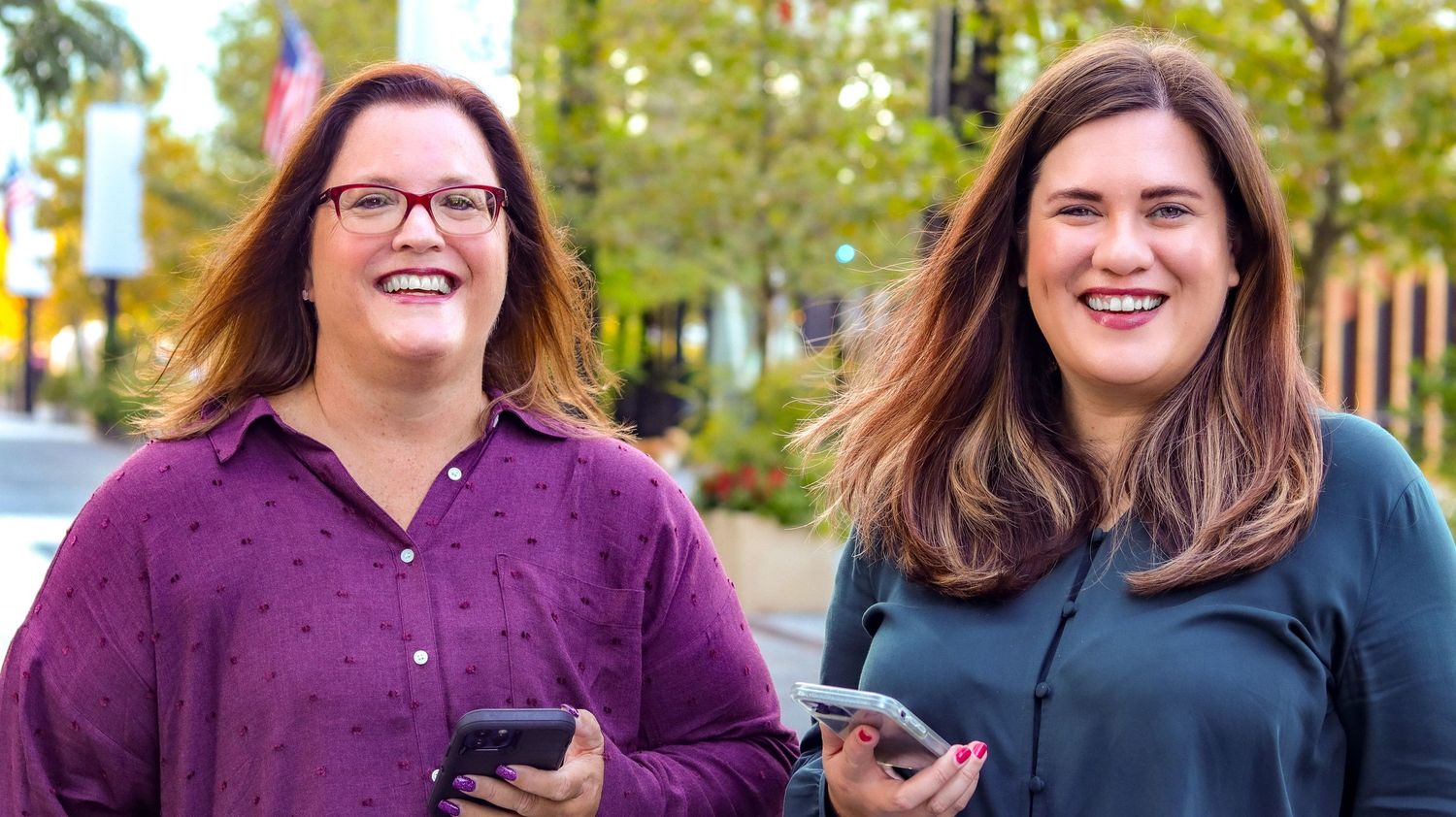 Two smiling women holding smartphones outdoors on a sunny day near colorful trees.