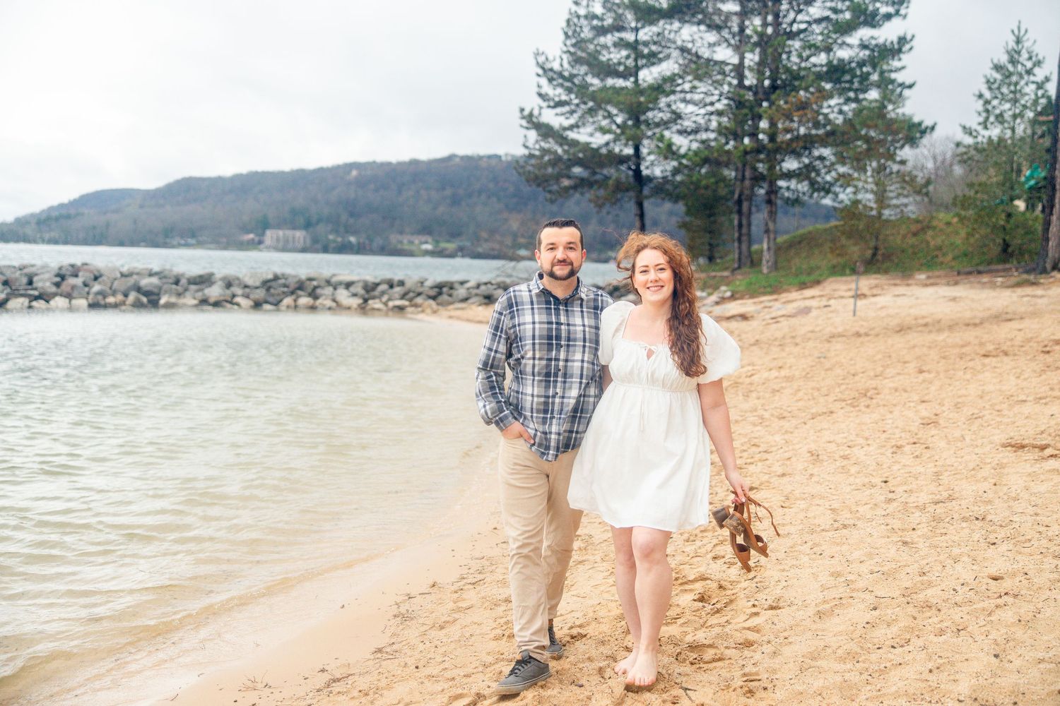Couple walking hand in hand on a sandy beach by a lake, surrounded by trees and hills in the background. deep creek lake state park md