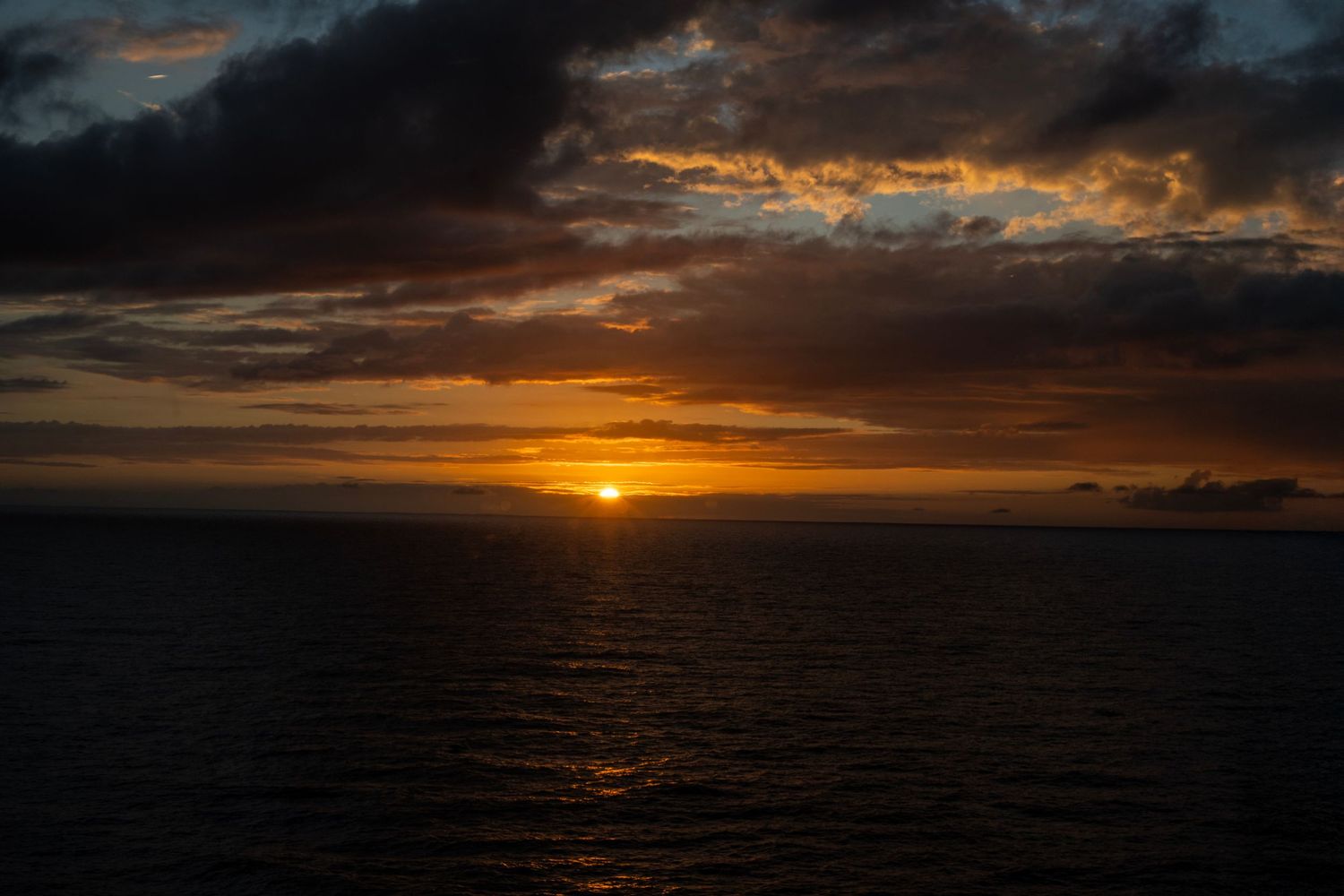 Dramatic ocean sunset with golden sun on horizon, dark storm clouds, and reflective water surface.