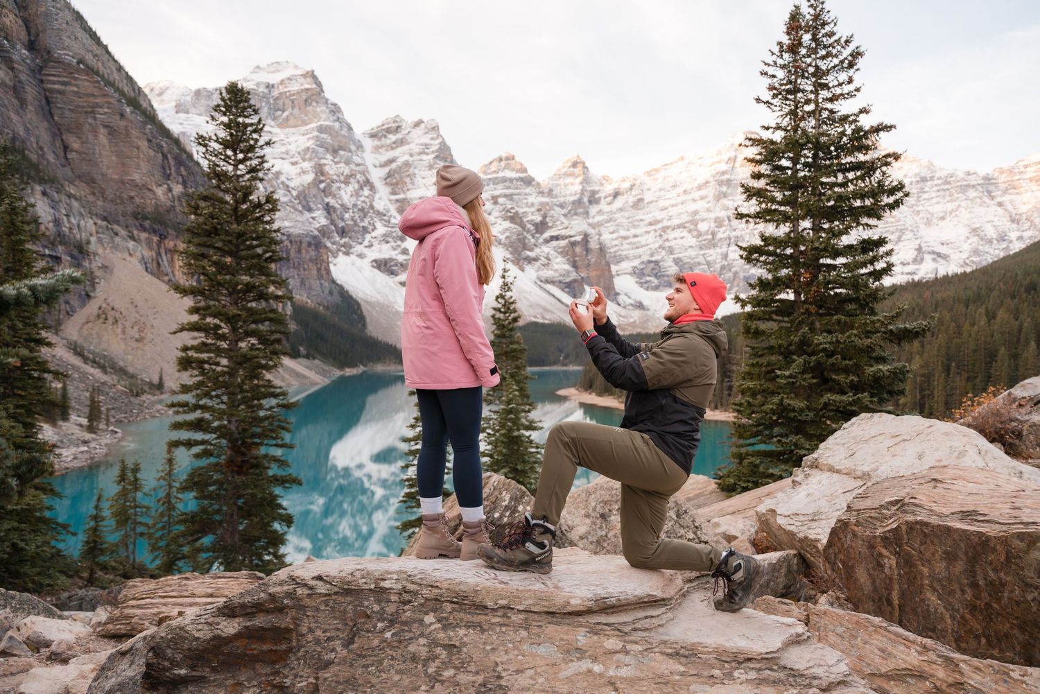 Man proposes to woman on rocky overlook at Moraine Lake, Banff, with turquoise water and snowy peaks behind them.