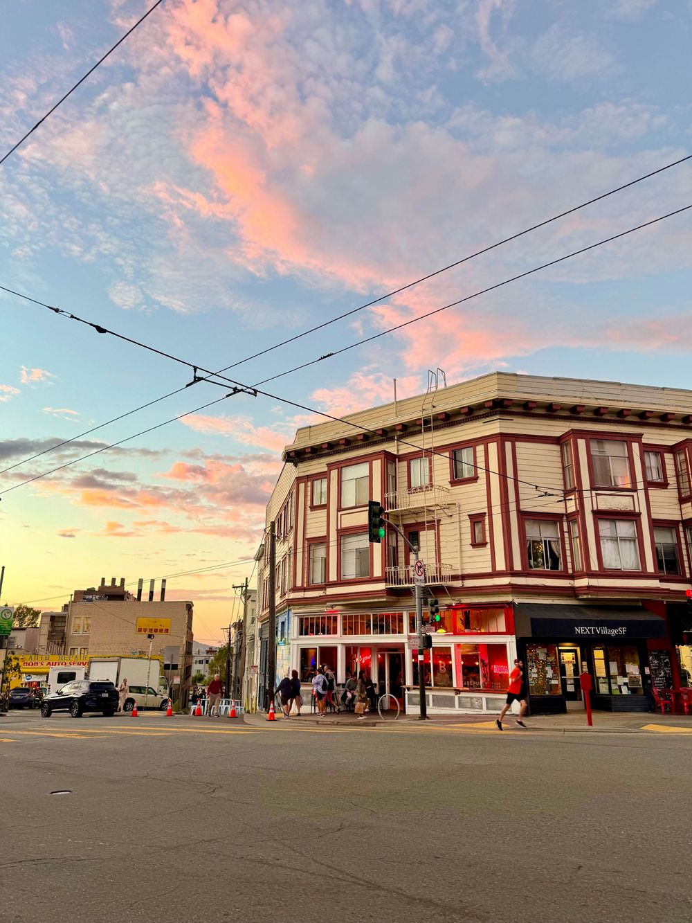 Colorful sunset sky over a historic corner building in San Francisco with trolley wires and busy street intersection.
