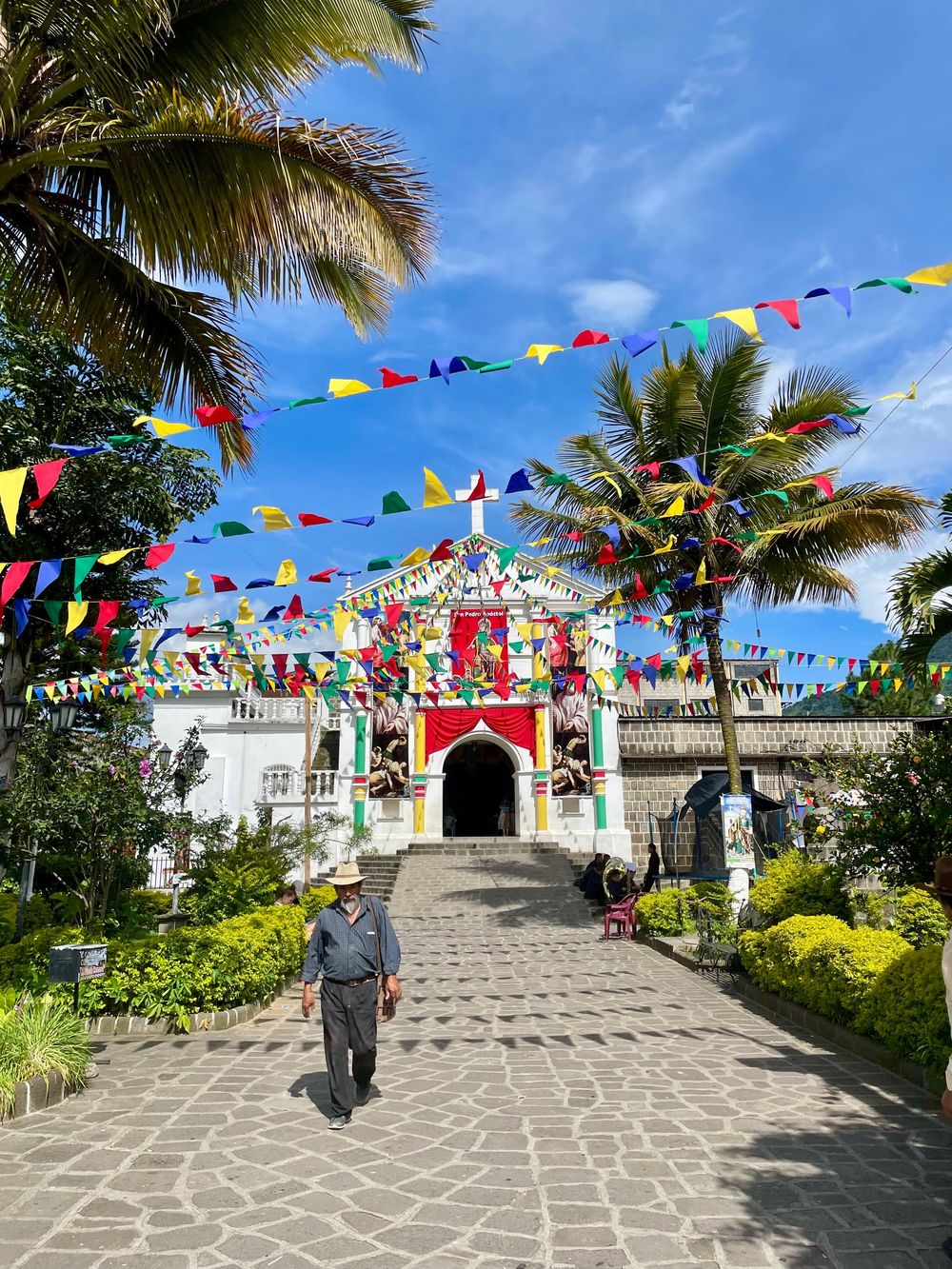 Colorful pennant flags decorate a festive white church entrance with palm trees under blue sky.