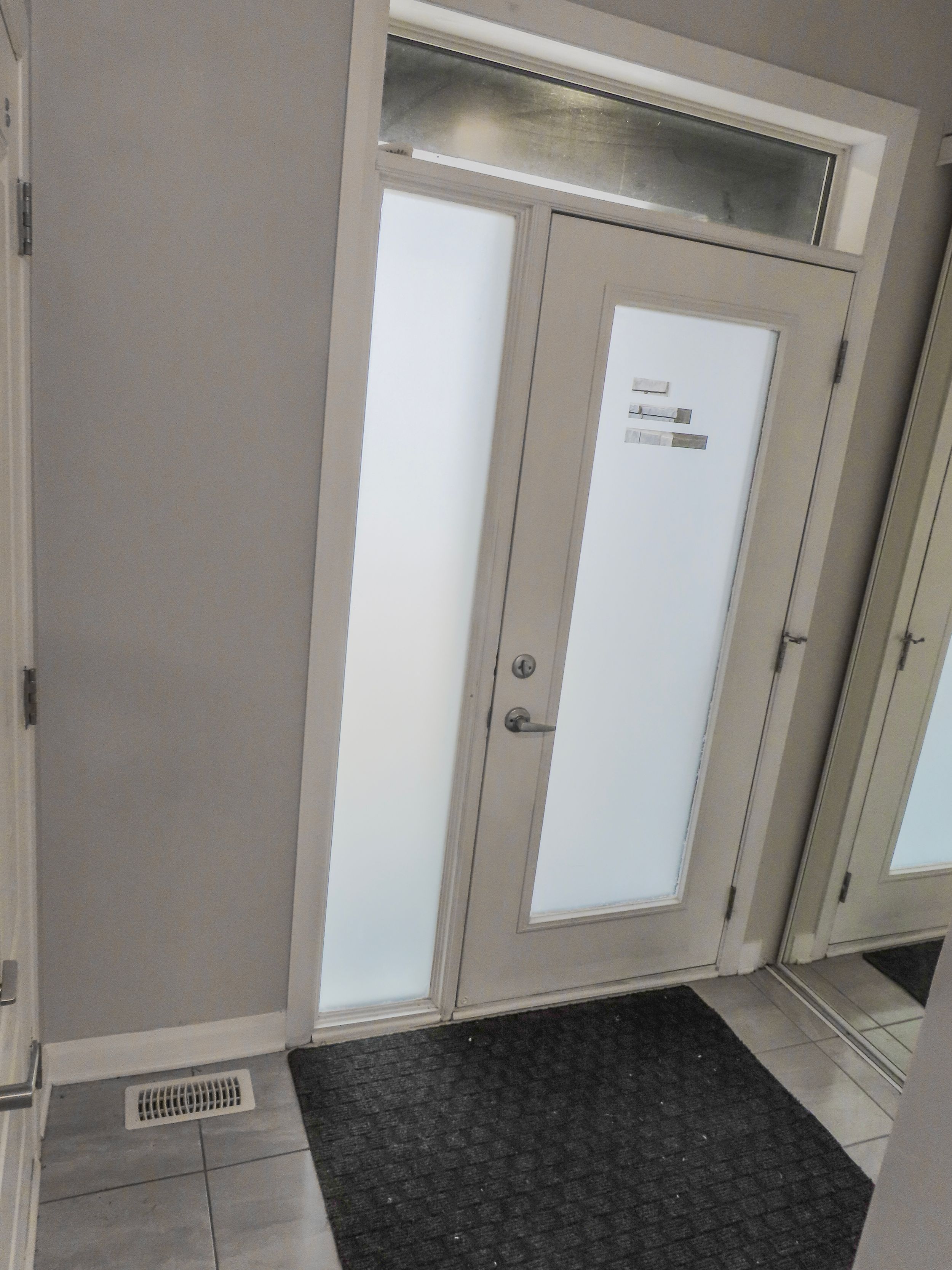 Modern front door with frosted glass panels, transom window, and sleek chrome hardware on white tile flooring.