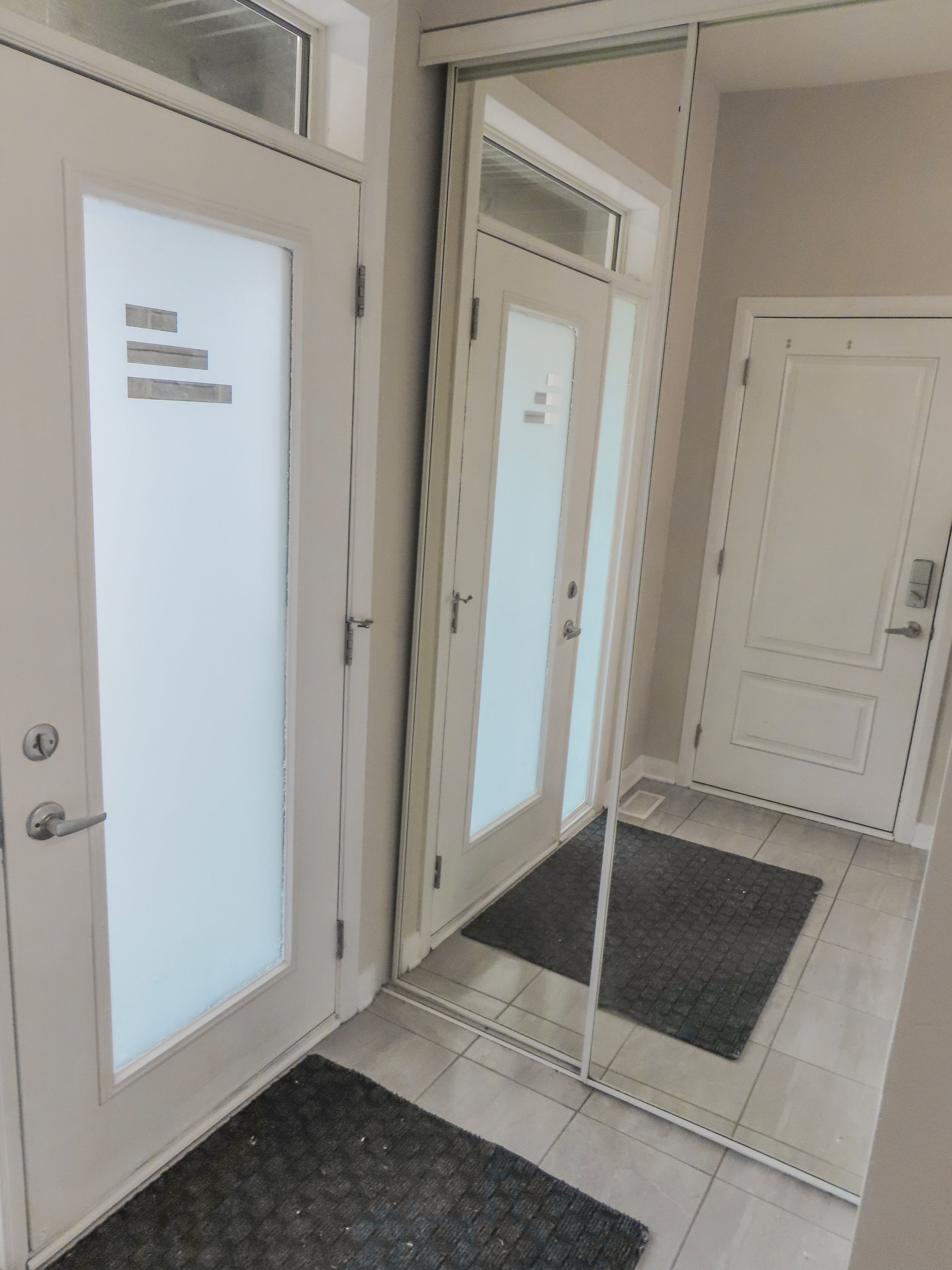 Modern entryway with frosted glass door, mirrored closet sliding doors, and tile flooring with dark welcome mat.