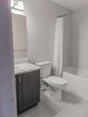 Modern bathroom with granite vanity, dark wood cabinets, white toilet, and tiled shower-tub combo with white curtain.