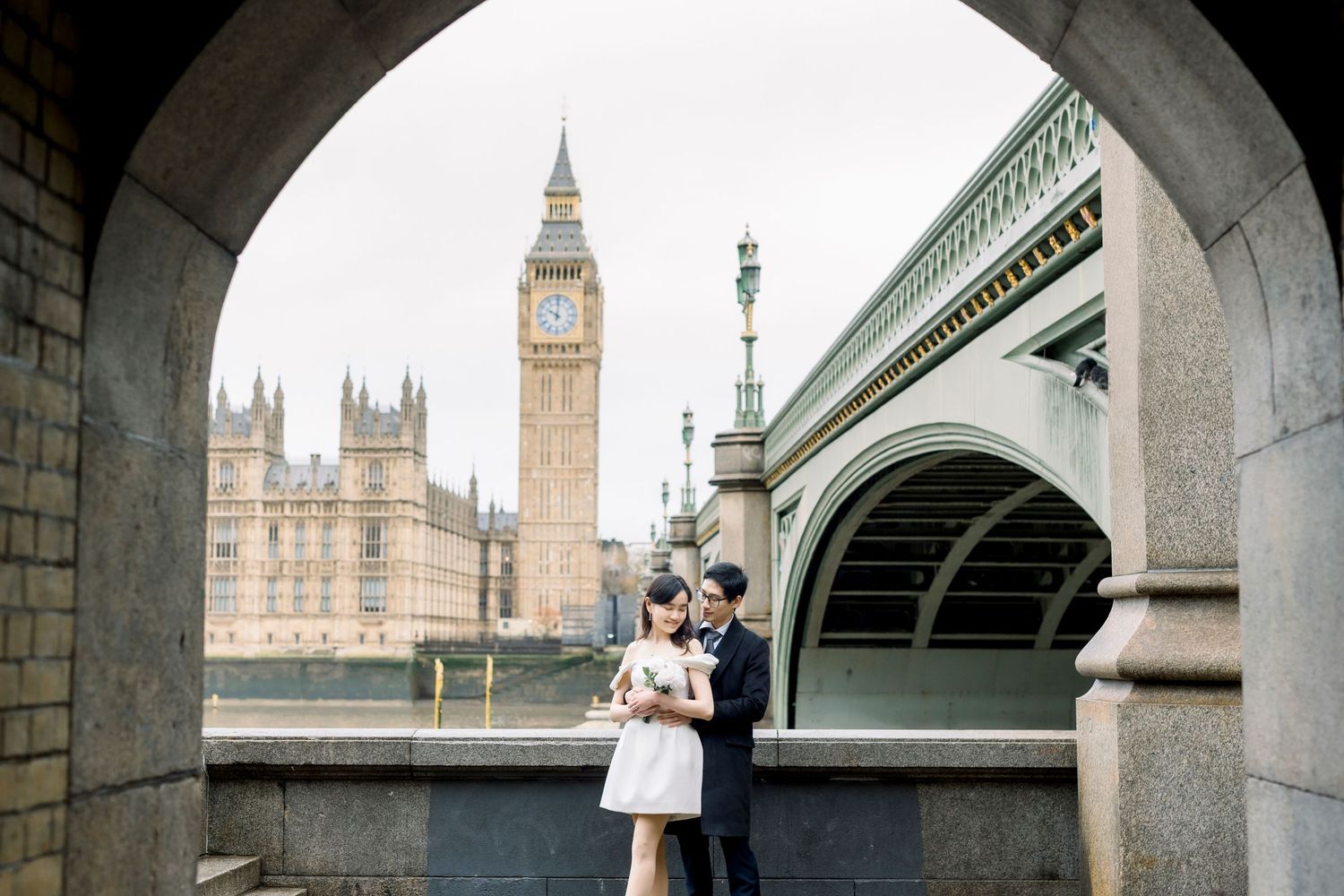 Couple embracing under arch with Big Ben and Westminster Bridge in background, London.