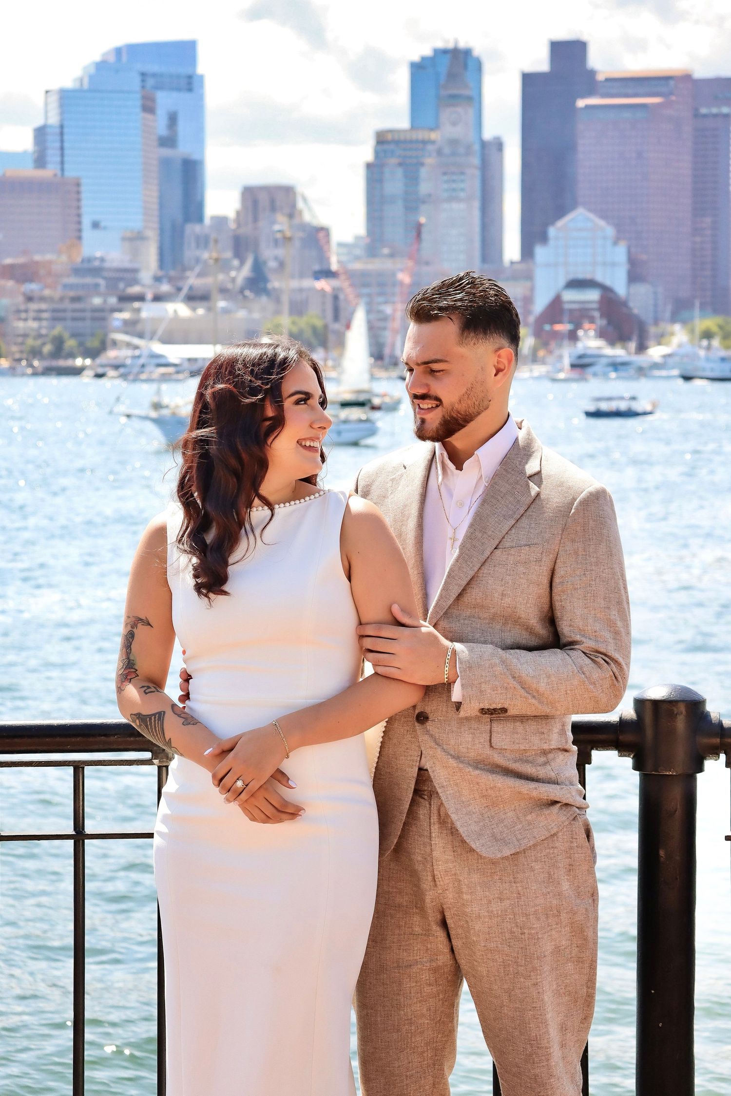 A couple poses along Boston Harbor with the city skyline in the background.
