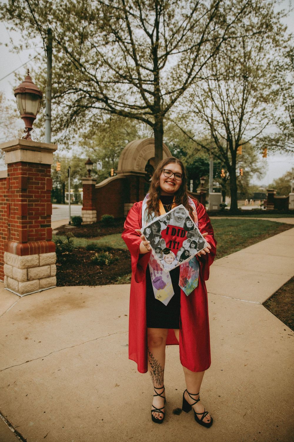 A smiling graduate in a red cap and gown holds a decorated mortarboard outdoors near a brick gate on a college campus.