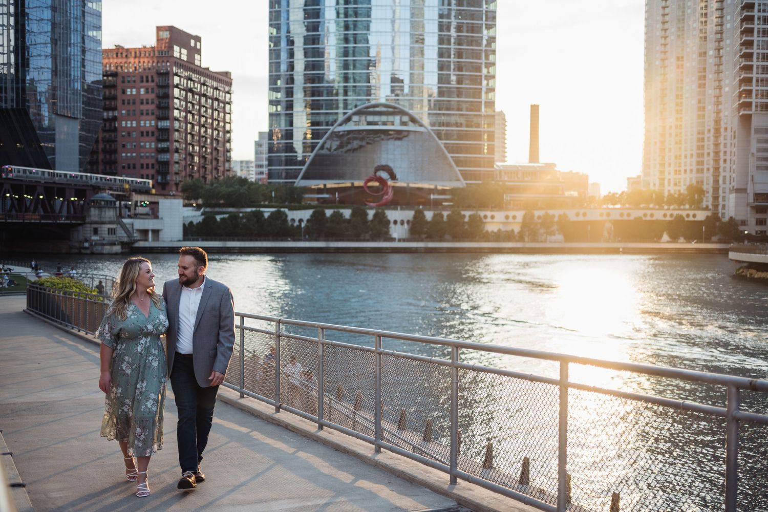 Couple walking along Chicago Riverwalk at golden hour with city skyline and glowing sunset in background.