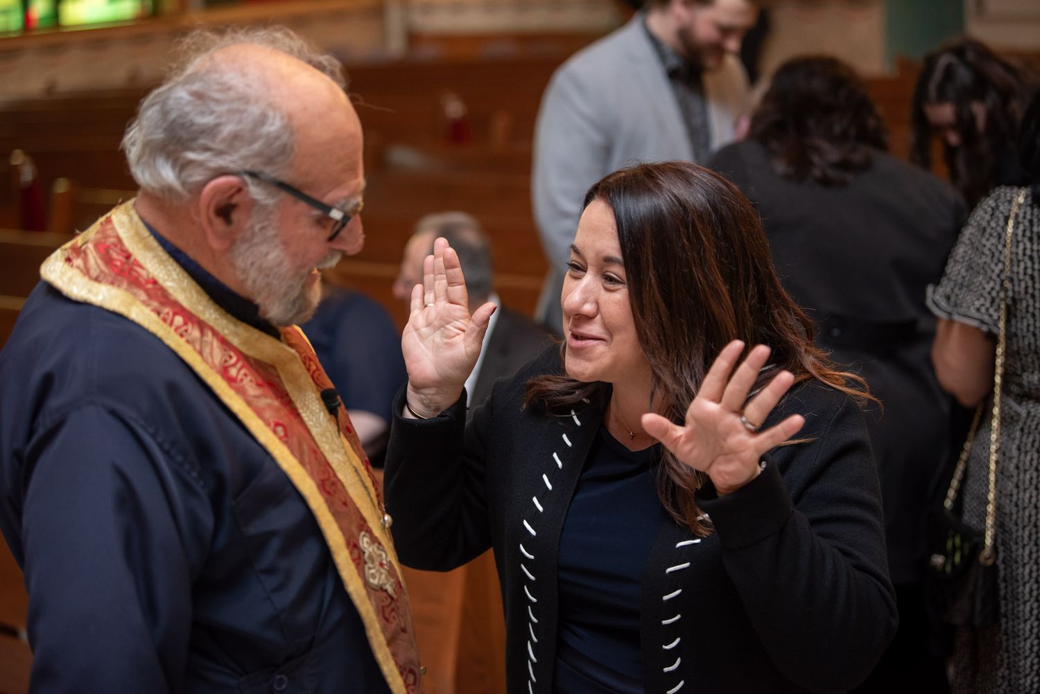 Orthodox priest in golden vestments speaking with young woman gesturing with hands in church.