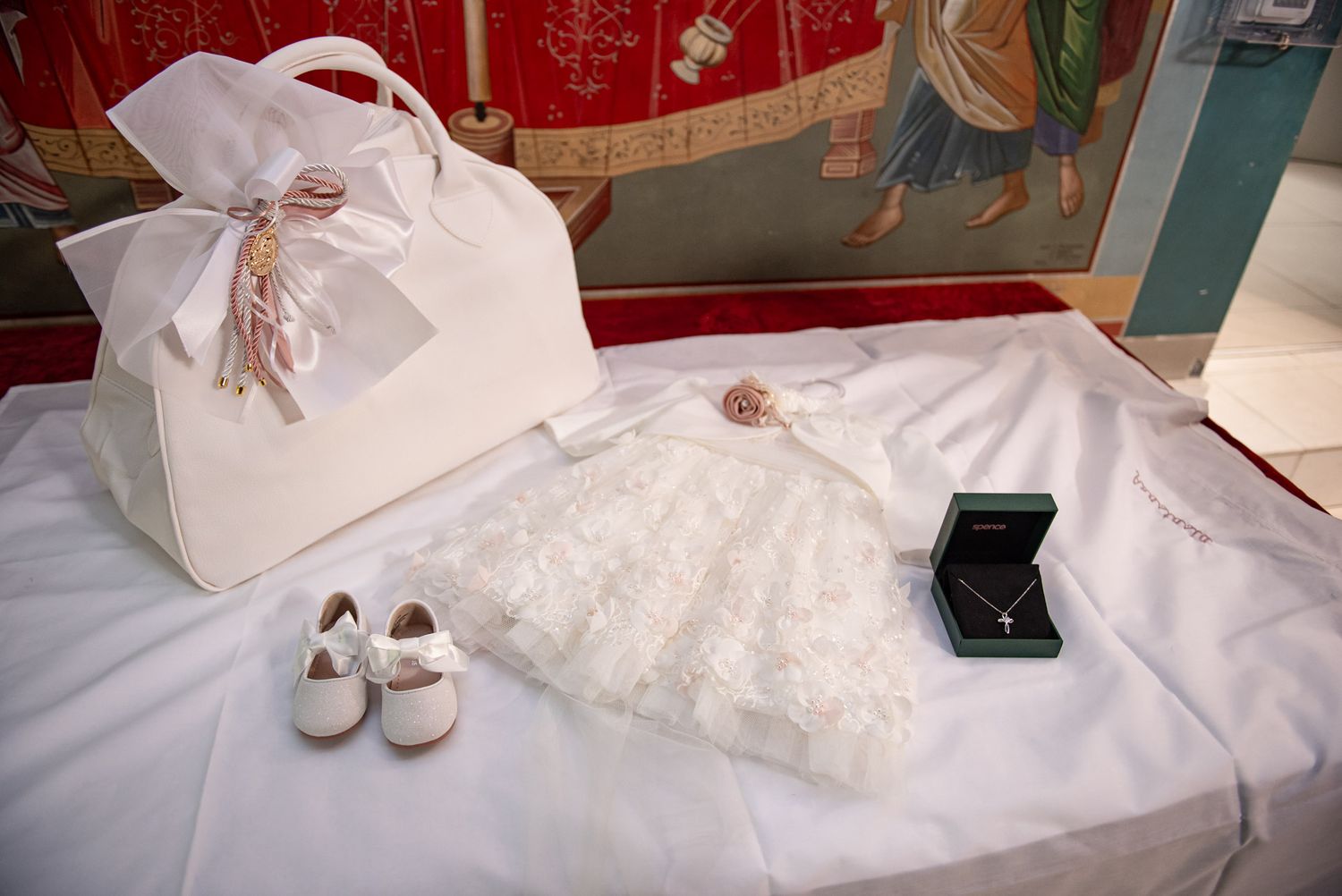White baptism accessories including lace dress, shoes, handbag, and jewelry box on white fabric.