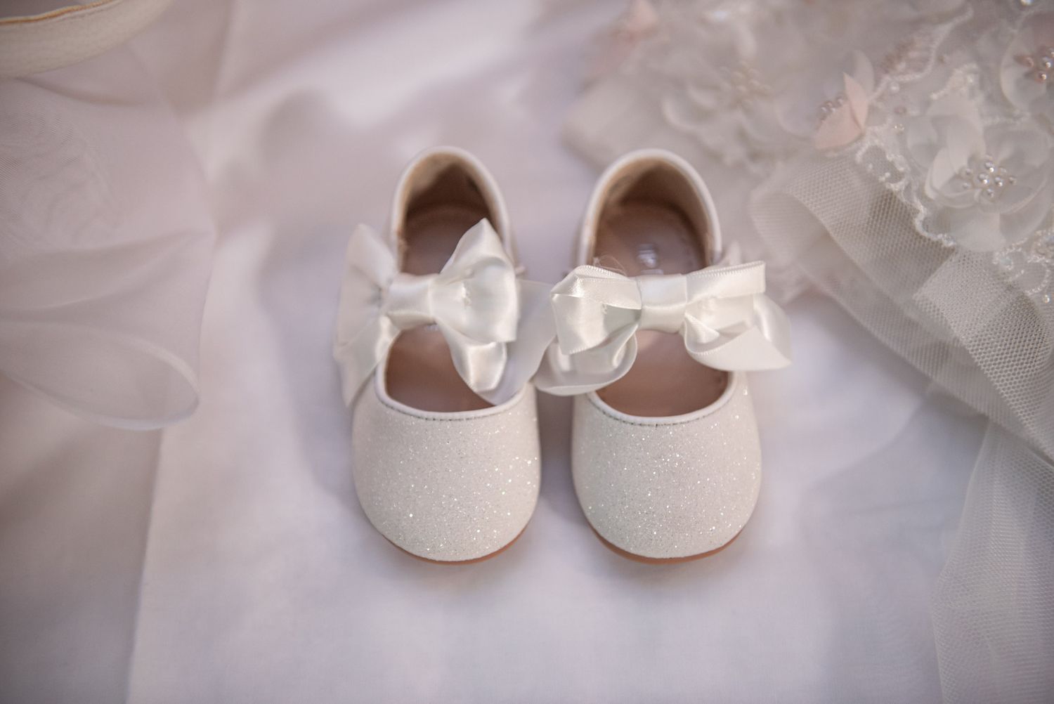 Pair of white glitter baby shoes with satin ribbon bows on white tulle fabric.