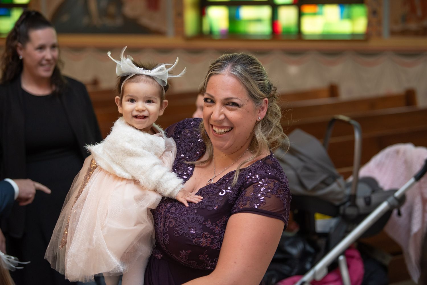 Woman in purple sequin dress holding smiling baby girl in white tutu outfit at church event.