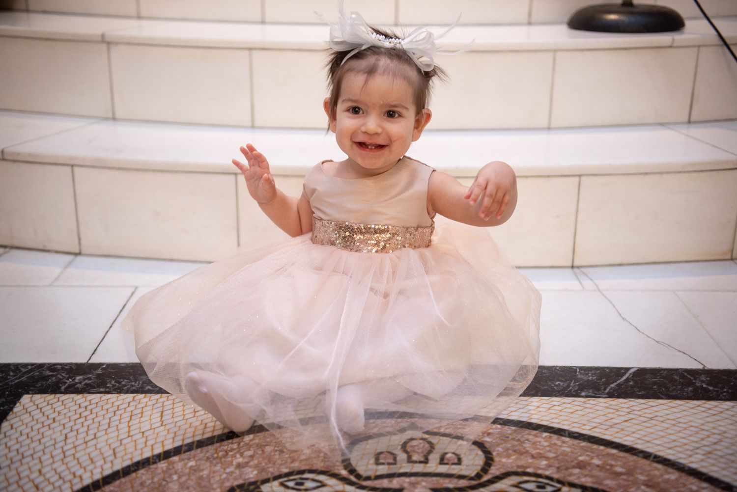 Happy baby girl in pink and gold tutu dress with bow headband sitting on marble steps.