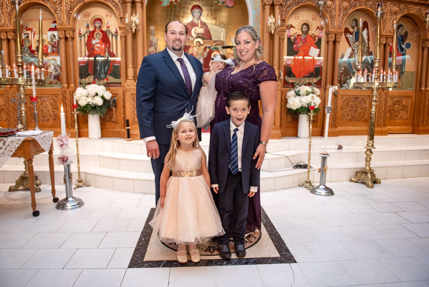 Family group portrait with godparents holding infant during Orthodox baptism ceremony at church altar.
