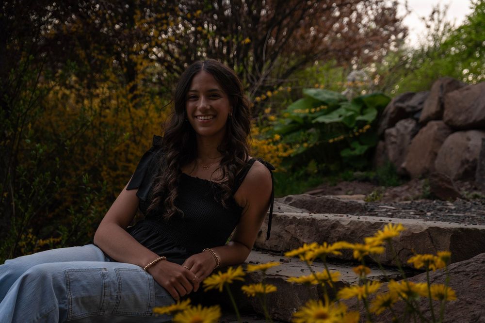 Young woman with long dark hair smiles while sitting outdoors among yellow flowers and lush greenery.