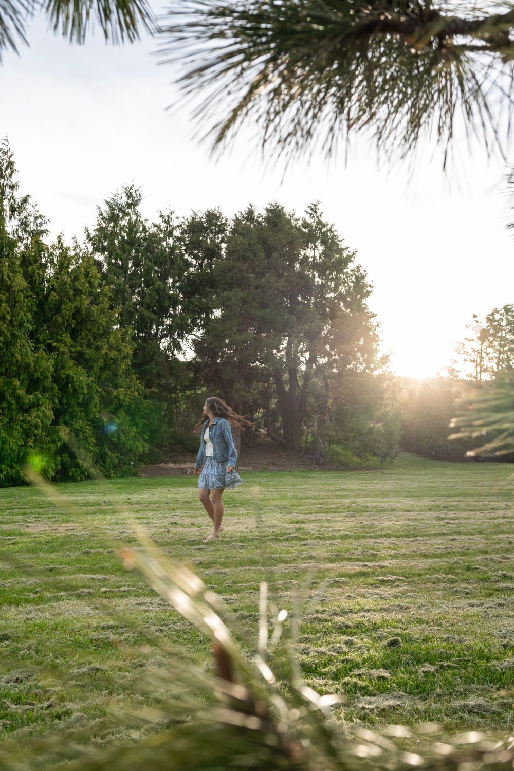 Woman walking on lush green grass in a sunlit park surrounded by trees during golden hour sunset.