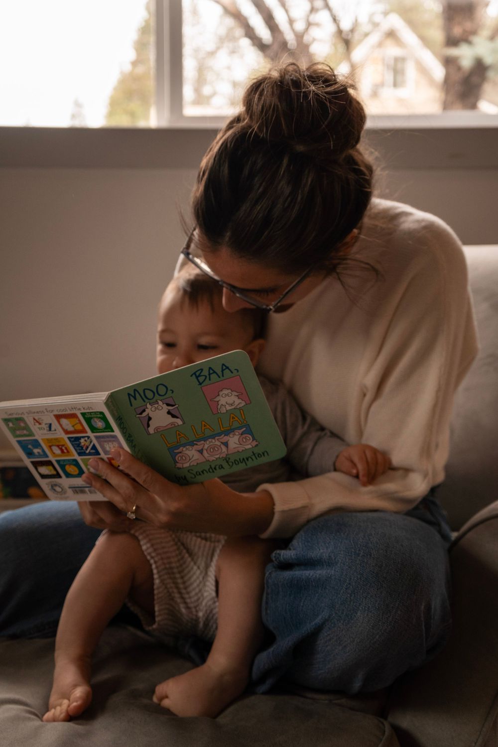 A mother reads the book Moo Baa La La La to her toddler on a couch near a bright window.