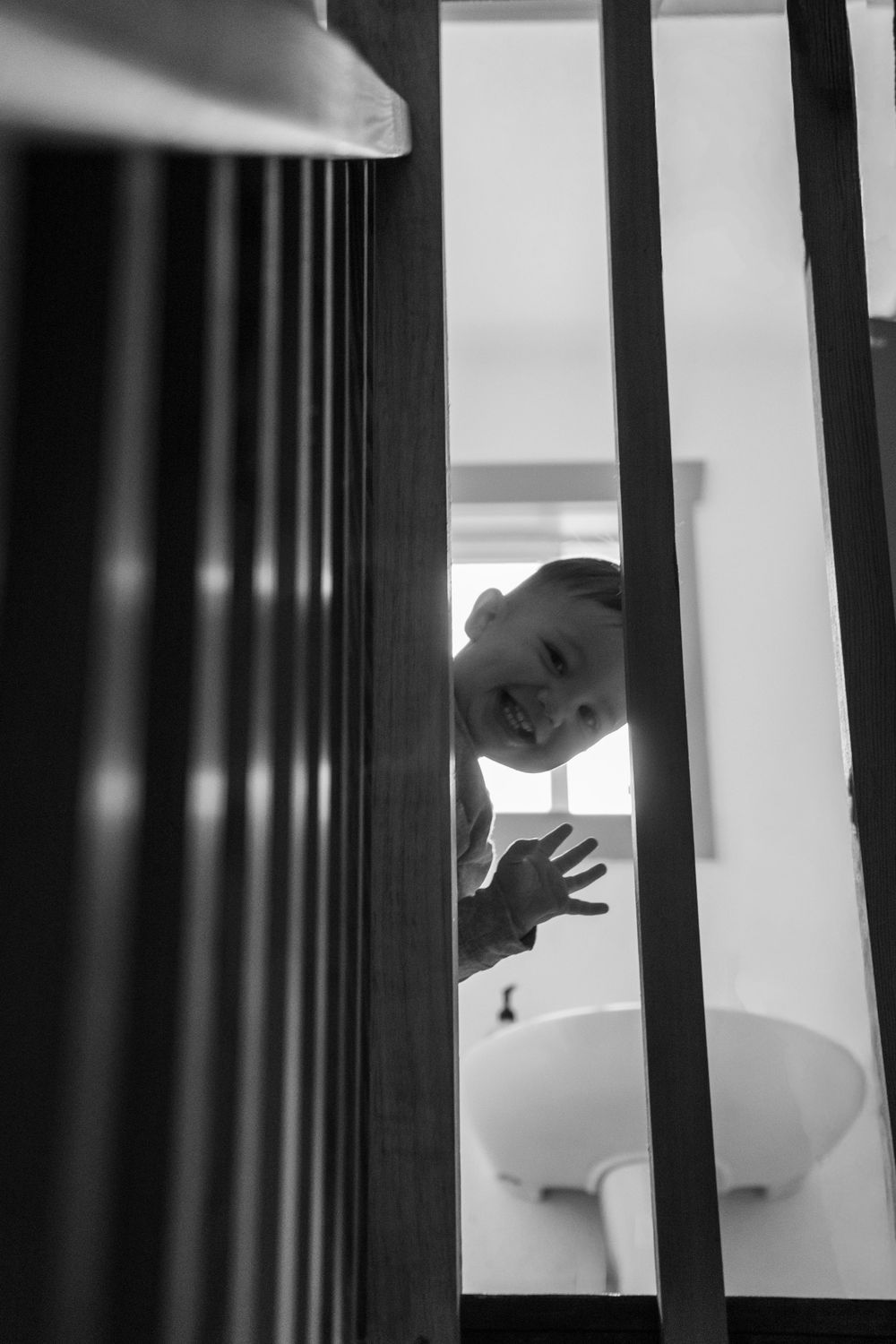 Black and white photo of a smiling child peeking through stair railings near a bathroom sink.