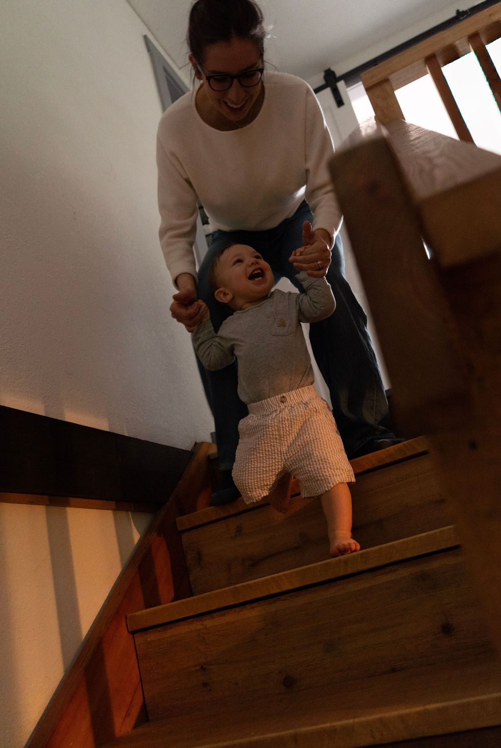 A smiling woman helps a happy toddler walk down wooden stairs inside a modern home.