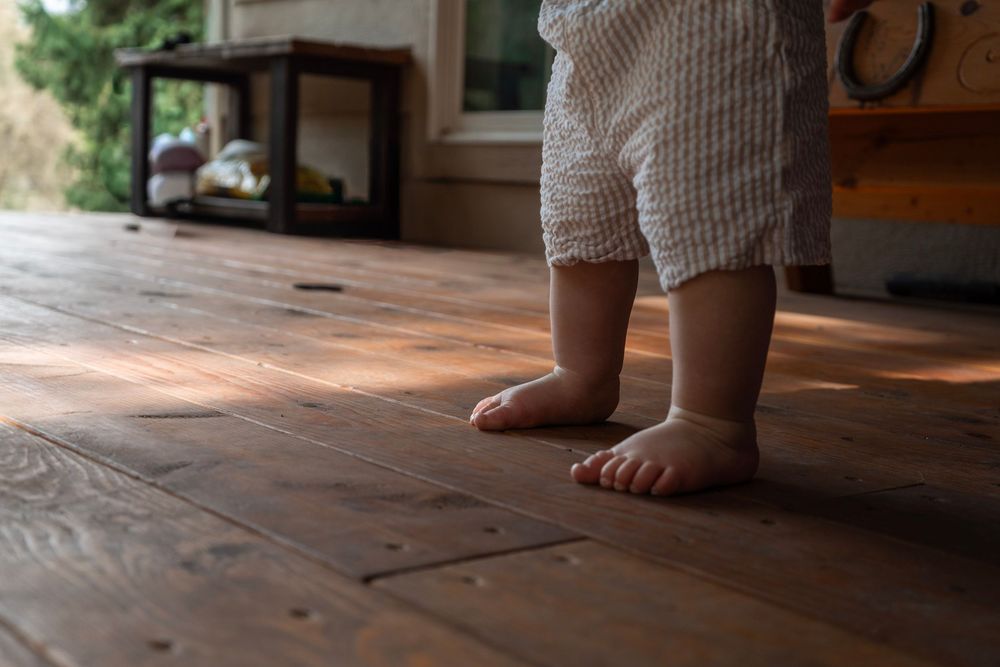 Close-up of baby's bare feet standing on rustic hardwood floor, taking first steps indoors.