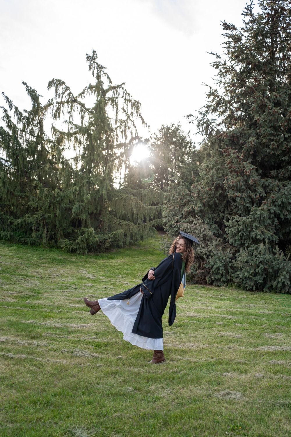 Young woman in graduation cap and gown twirling outdoors on green grass with tall evergreen trees and sunlight behind her.