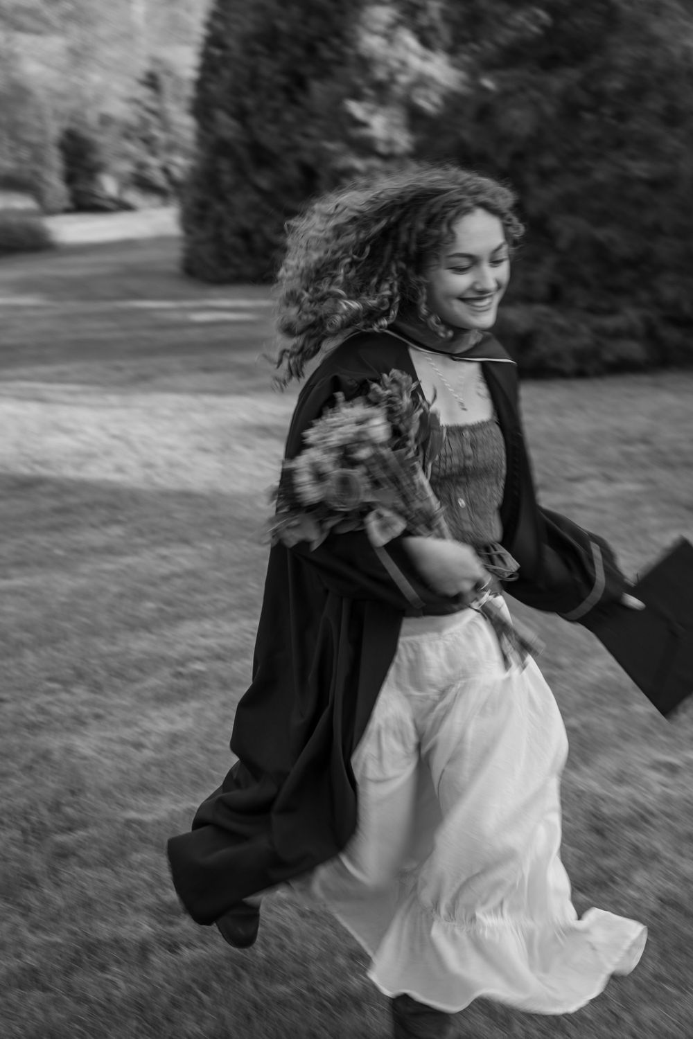 Black and white photo of a smiling woman with curly hair running outdoors in a flowing skirt and jacket.