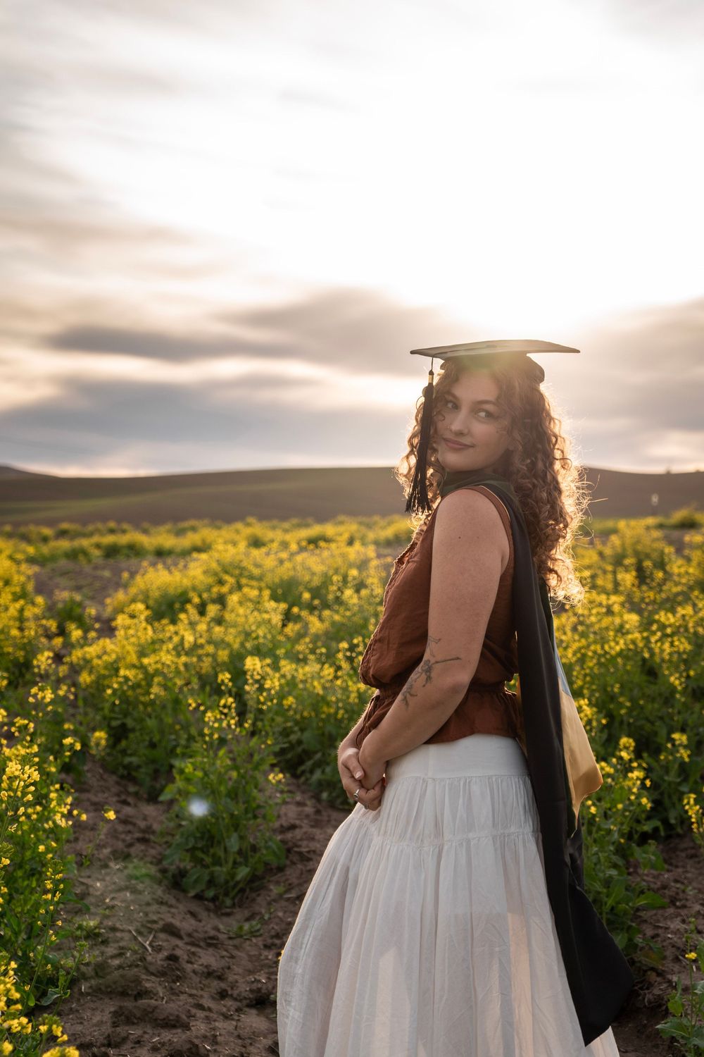 Curly-haired woman in graduation cap standing in yellow flower field at golden sunset.