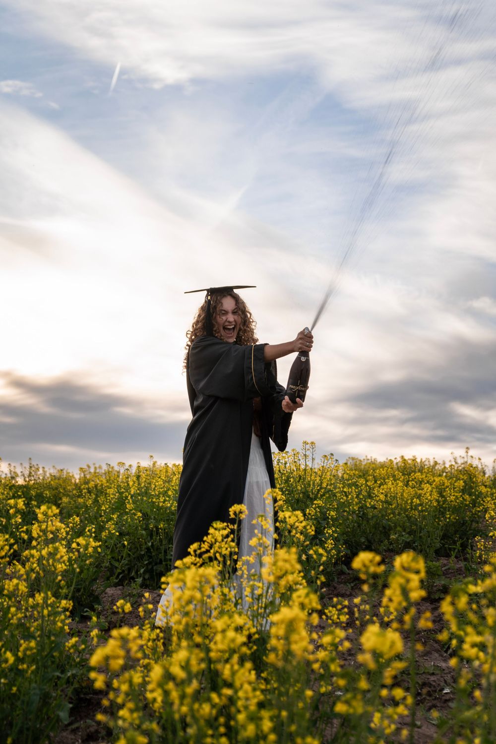 Graduate in black cap and gown wielding a sword in a yellow flower field at sunset.