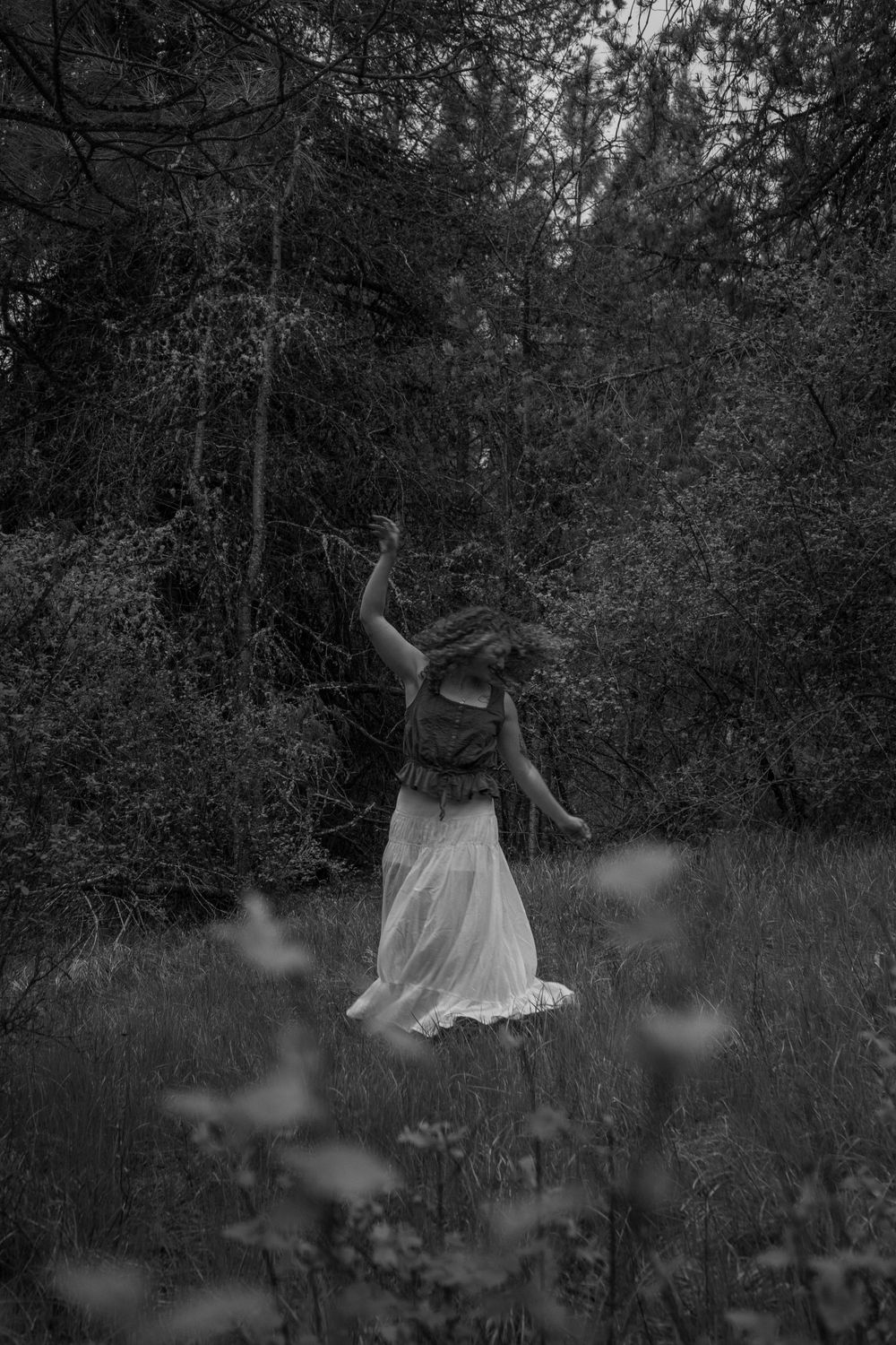 Black and white photo of a woman in a white dress dancing freely in a dark, mystical forest clearing.