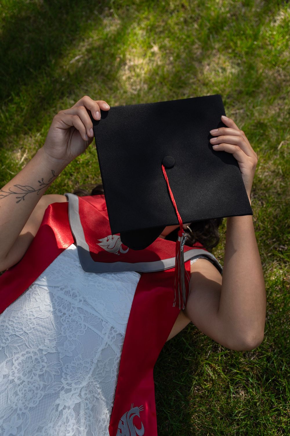 Graduate lying on grass holding black mortarboard cap, wearing red WSU graduation gown with white stole.