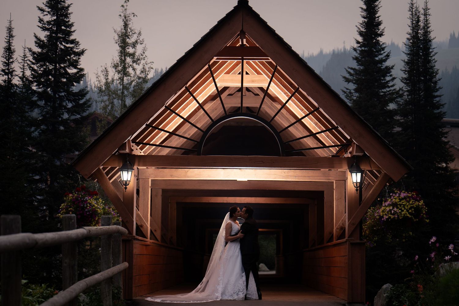 Silhouetted wedding couple stands in illuminated wooden chapel entrance at dusk.