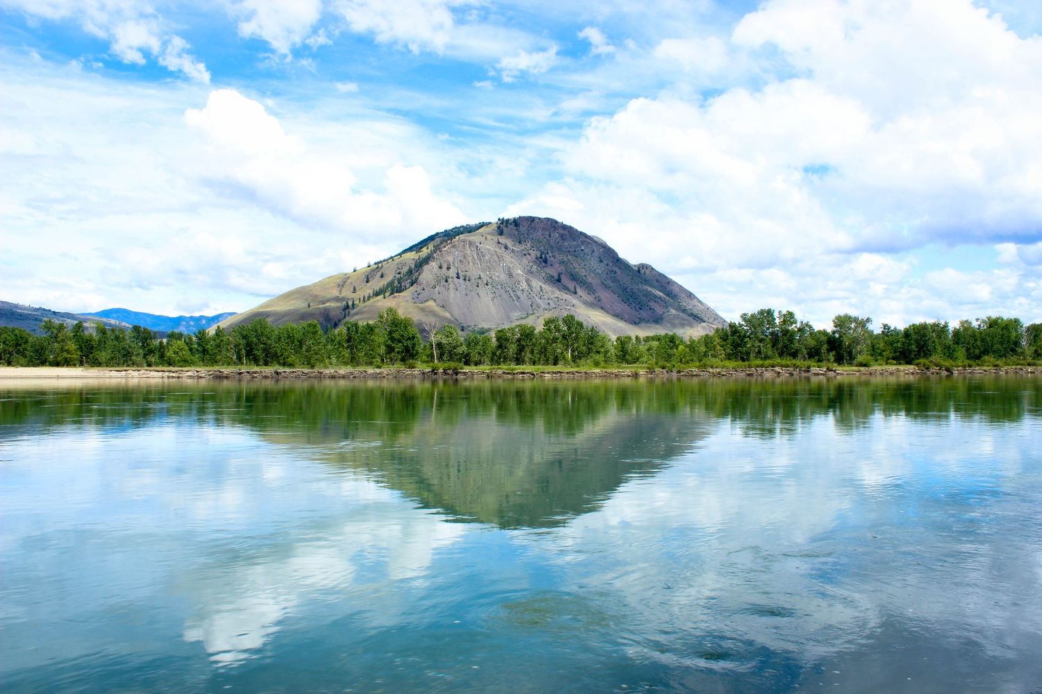Serene mountain lake reflecting blue skies and green trees with a majestic peak in the background.