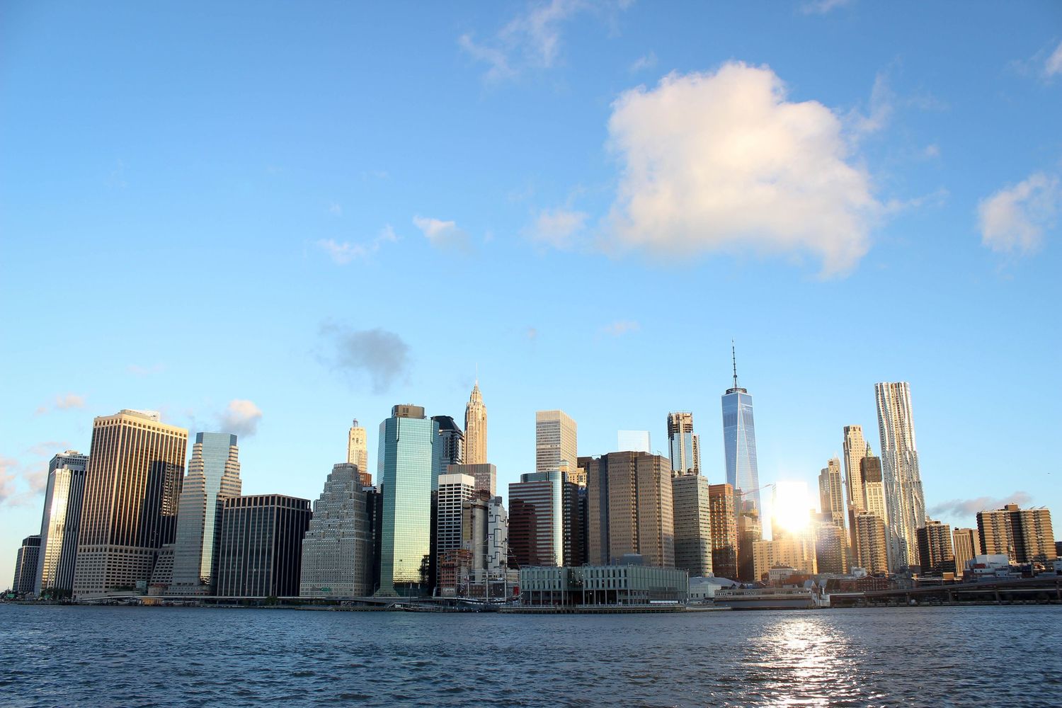 Manhattan skyline with modern skyscrapers reflecting sunset light across the harbor.