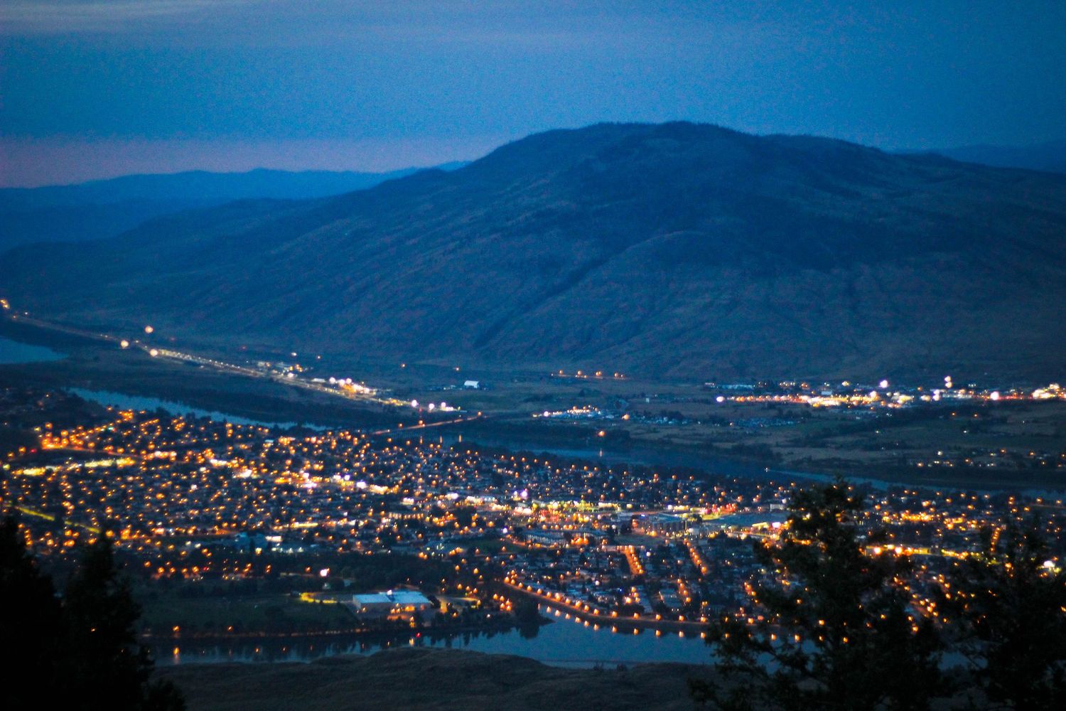 Aerial cityscape at dusk with mountains and twinkling lights reflecting across the urban landscape.