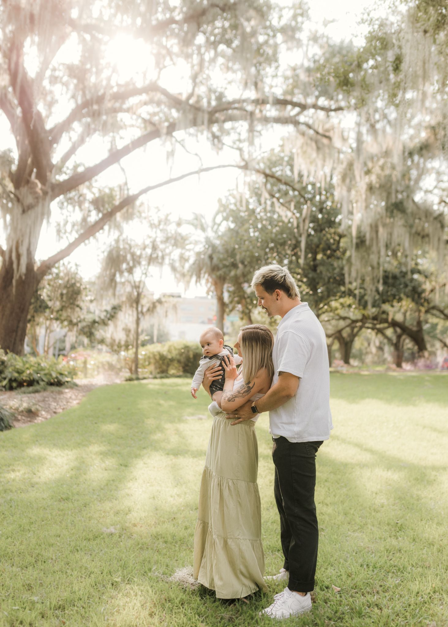 A young family with a baby poses under Spanish moss-draped oak trees in a sunlit park setting.