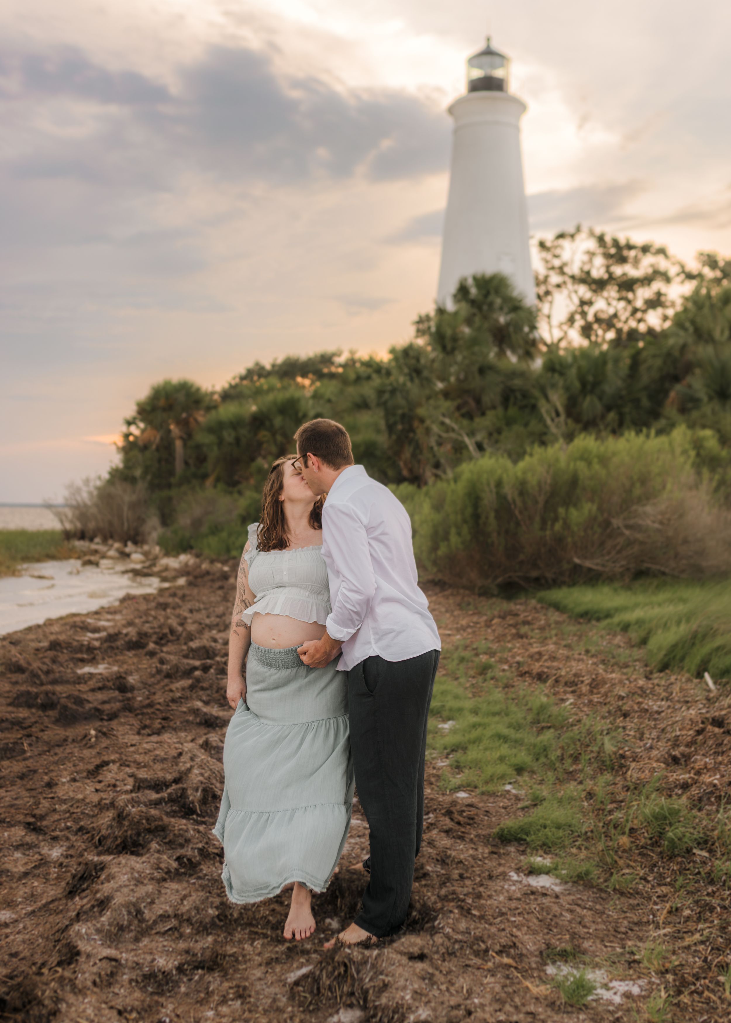 Pregnant couple sharing a kiss at sunset near a white lighthouse, surrounded by lush greenery and coastal scenery.