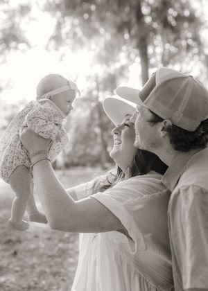 Black and white photo of parents joyfully lifting their smiling baby outdoors among trees.
