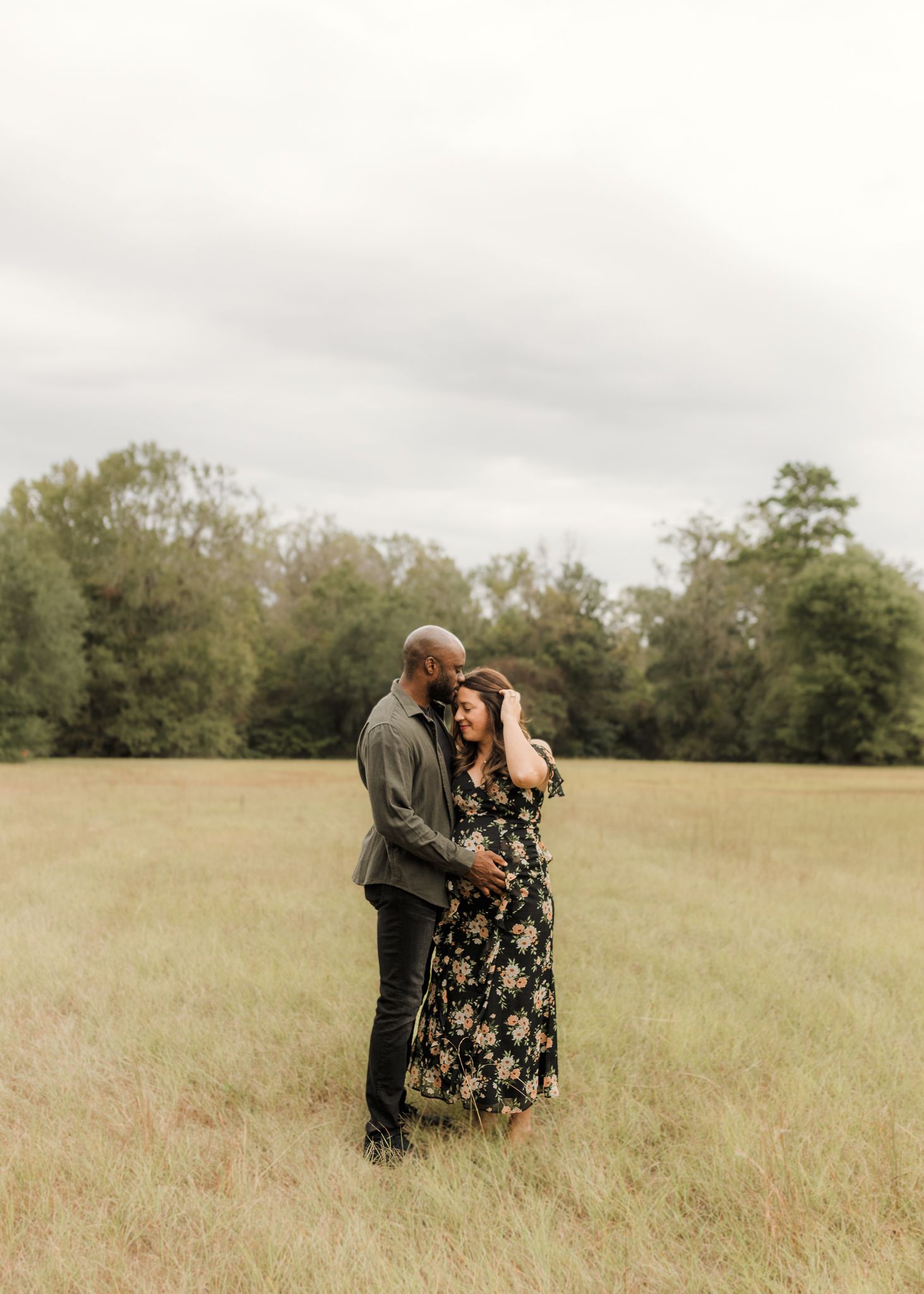 Couple embracing romantically in an open grassy field with lush green trees and overcast sky backdrop.