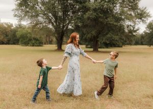 A red-haired woman in a floral dress holds hands with two young boys in a green field.