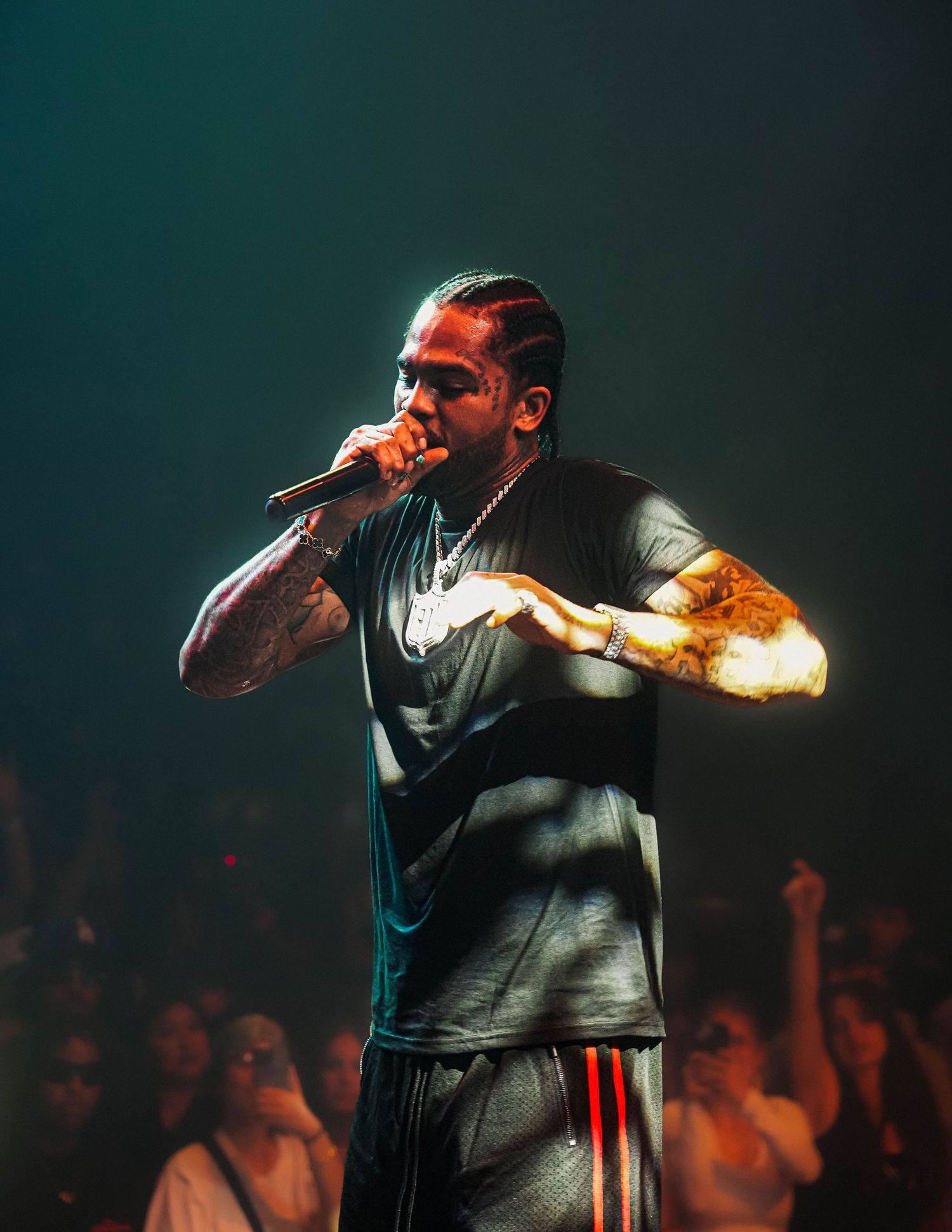 A performer in a dark t-shirt energetically raps on stage in dramatic lighting while a crowd cheers in the background.
