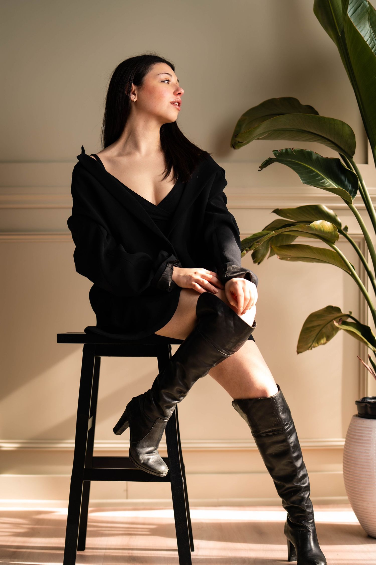 Fashion portrait of woman in black sweater and tall boots sitting on stool by houseplant.
