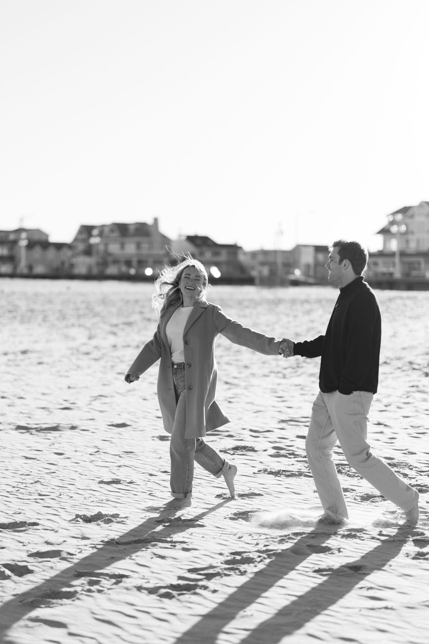 Black and white photo of a happy couple holding hands and running on a sandy beach with houses in the background.