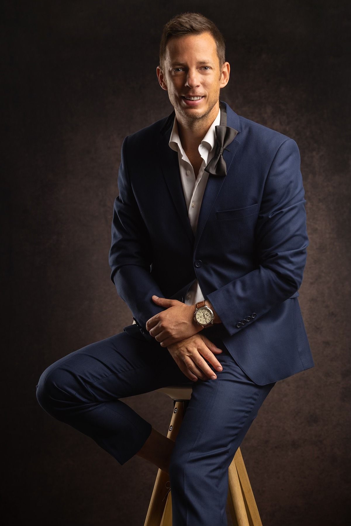 Smiling man in navy blue suit sitting on wooden stool against dark background, wearing watch.