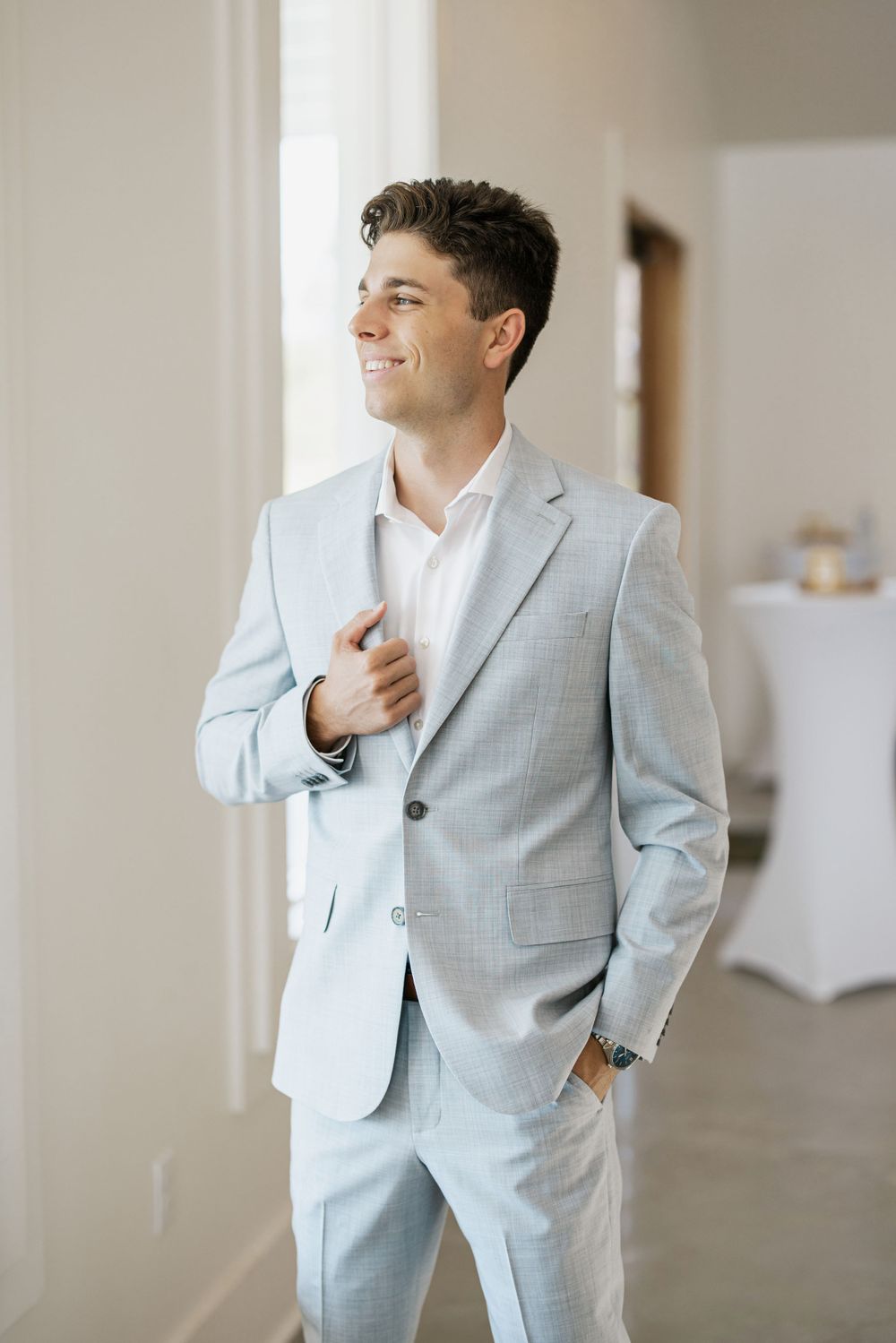 A groom in a light blue suit stands confidently in an elegant indoor setting.