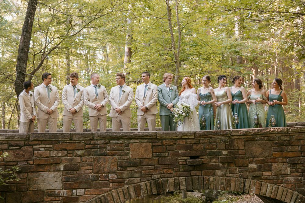 Wedding party in pastel attire poses together on a rustic stone bridge in a forest setting.
