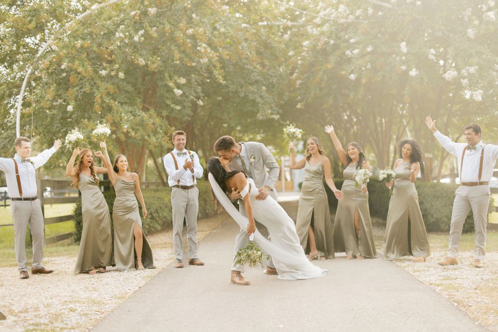 Wedding party celebrates with dip pose on path surrounded by trees in golden sunlight.