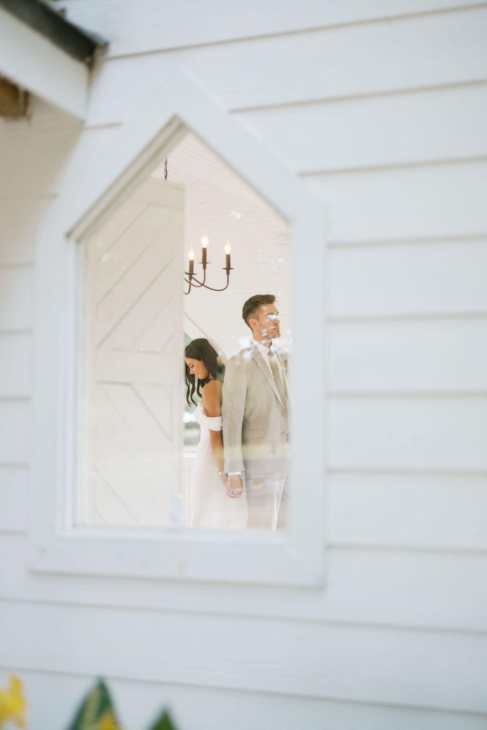A couple peeks through a white church window frame during their wedding photoshoot.